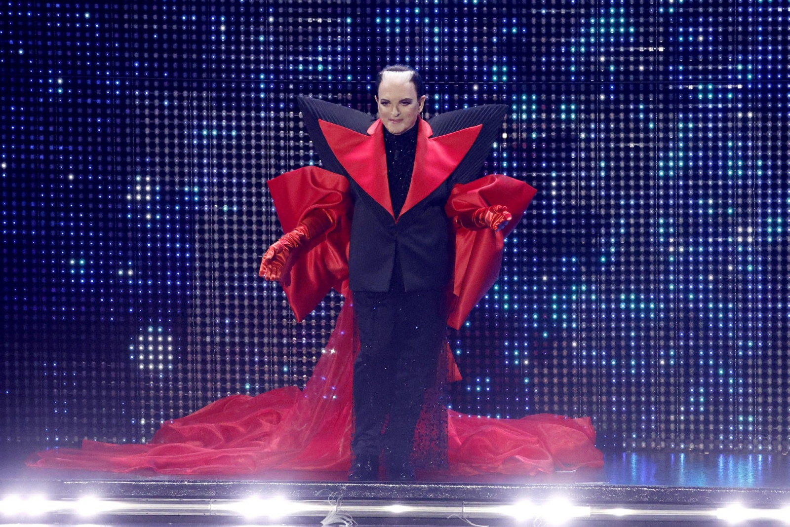 Abituato a stupire con i suoi look stravaganti, per il suo debutto sul palco di Sanremo Cristiano Malgioglio ha scelto un appariscente abito nero e rosso, con enorme bavero scultura e strascico di velo rosso 